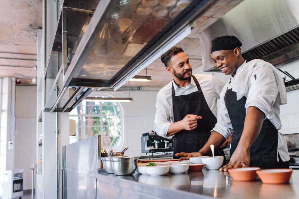 Happy men chefs cooking food at cafe kitchen. Two cooks making food dishes in restaurant kitchen.